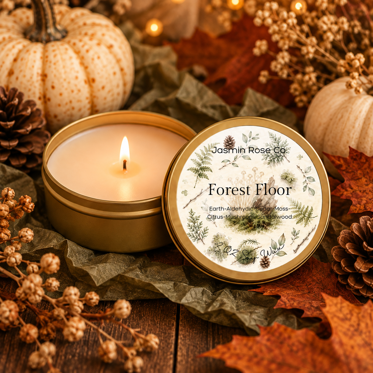 A lit candle in a round brass tin sits beside its matching lid on a rustic wooden surface, surrounded by dried leaves, pinecones, small pumpkins, and warm string lights. The lid label reads 'Jasmin Rose Co' and 'Forest Floor'.