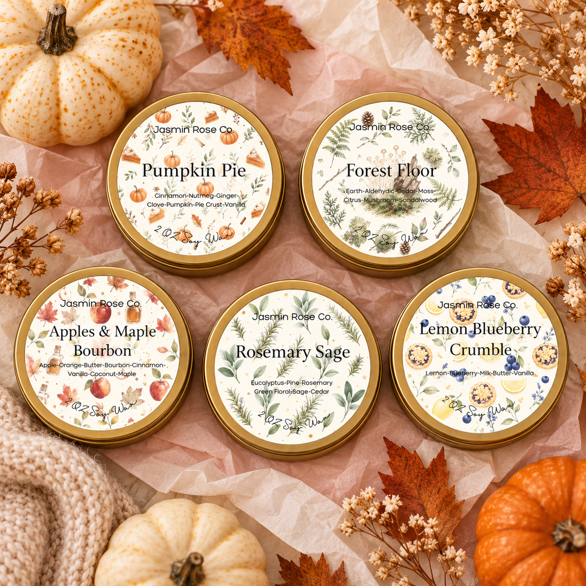 Five round fall scented candles in gold-rimmed tins from Jasmine Rose Co., arranged on pink tissue paper with autumn-themed props including pumpkins, dried flowers, and leaves.