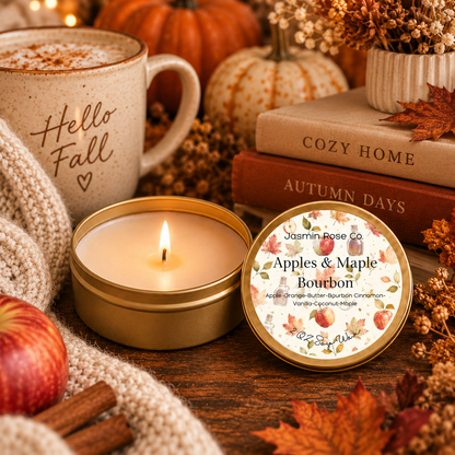 A lit candle in a round gold tin sits beside its printed lid on a rustic wooden table, surrounded by fall-themed props including a mug reading 'Hello Fall,' stacked books, cinnamon sticks, an apple, dried leaves, and pumpkins. The lid shows the brand Jasmin Rose Co and the scent name Apples & Maple Bourbon.