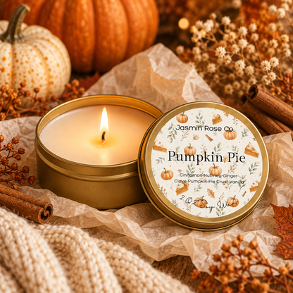 A lit candle in a round gold tin sits on parchment paper with its lid beside it. The lid label reads 'Jasmin Rose Co' and 'Pumpkin Pie,' with autumn-themed illustrations.